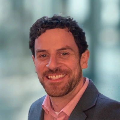 Portrait of Dr. Daniel Buckley wearing a pink shirt and a gray blazer, sporting a big smile.